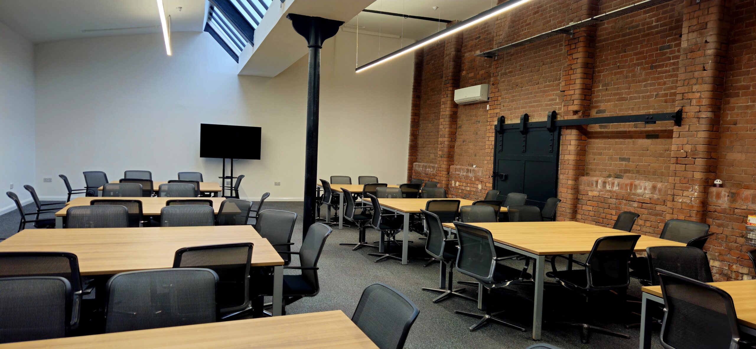 stylish meeting room at Ram Mill Business Centre in Oldham, featuring exposed brick walls, natural light from a skylight, modern furniture, and a large screen for presentations. Perfect for business meetings, workshops, and training sessions