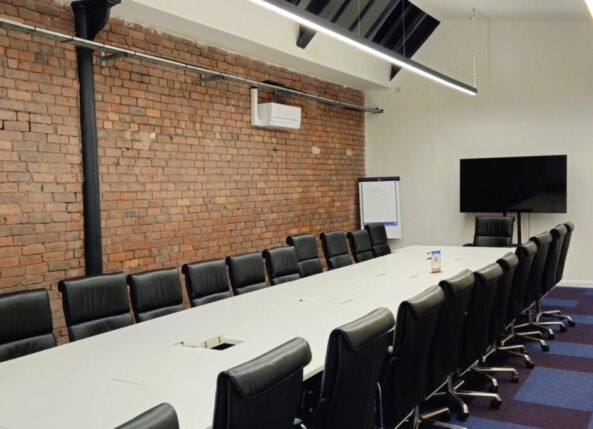 xecutive-style meeting room at Ram Mill Business Centre in Oldham, featuring a long conference table, sleek leather chairs, exposed brick walls, and a large screen for presentations. Ideal for board meetings, client pitches, and professional gatherings