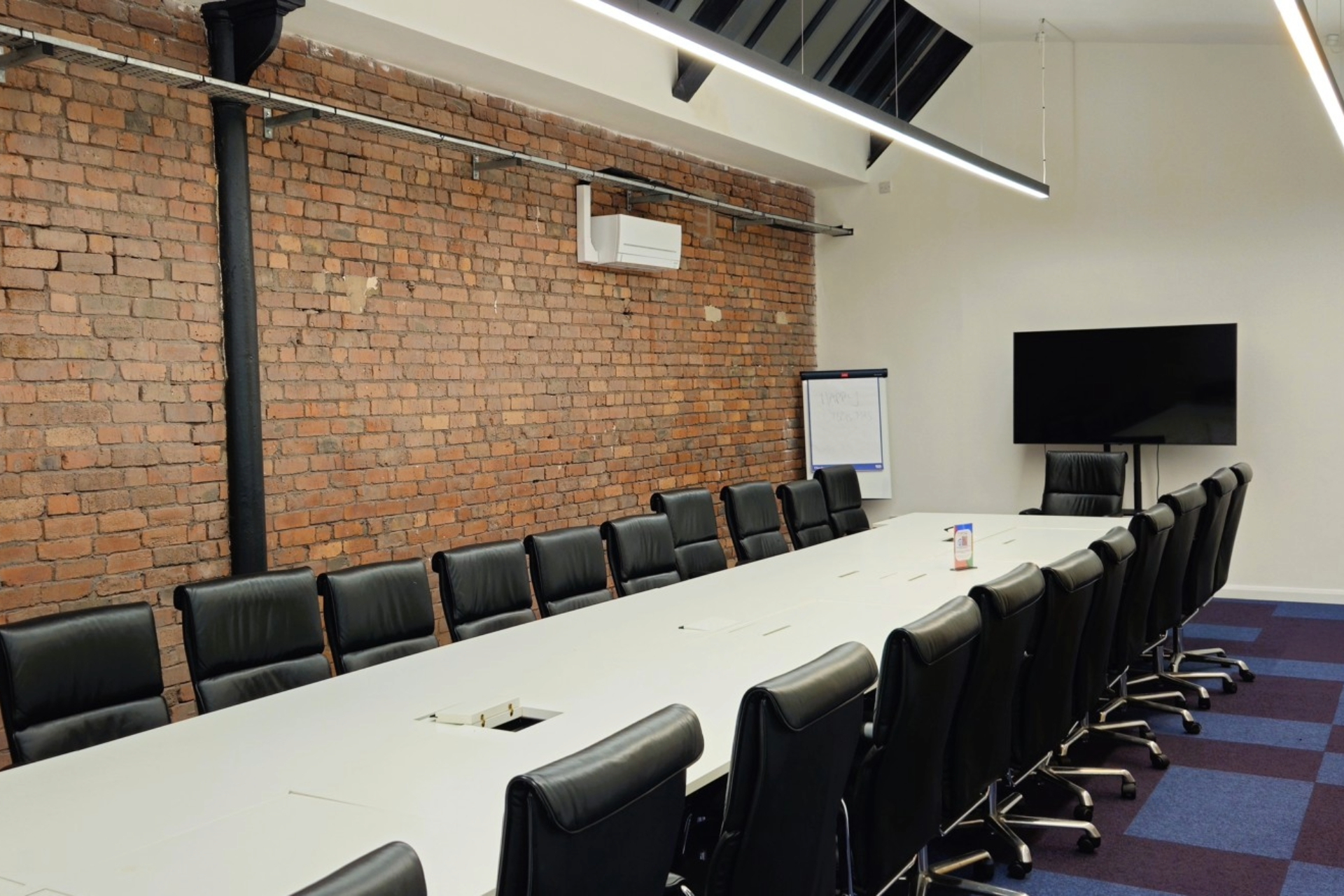 xecutive-style meeting room at Ram Mill Business Centre in Oldham, featuring a long conference table, sleek leather chairs, exposed brick walls, and a large screen for presentations. Ideal for board meetings, client pitches, and professional gatherings