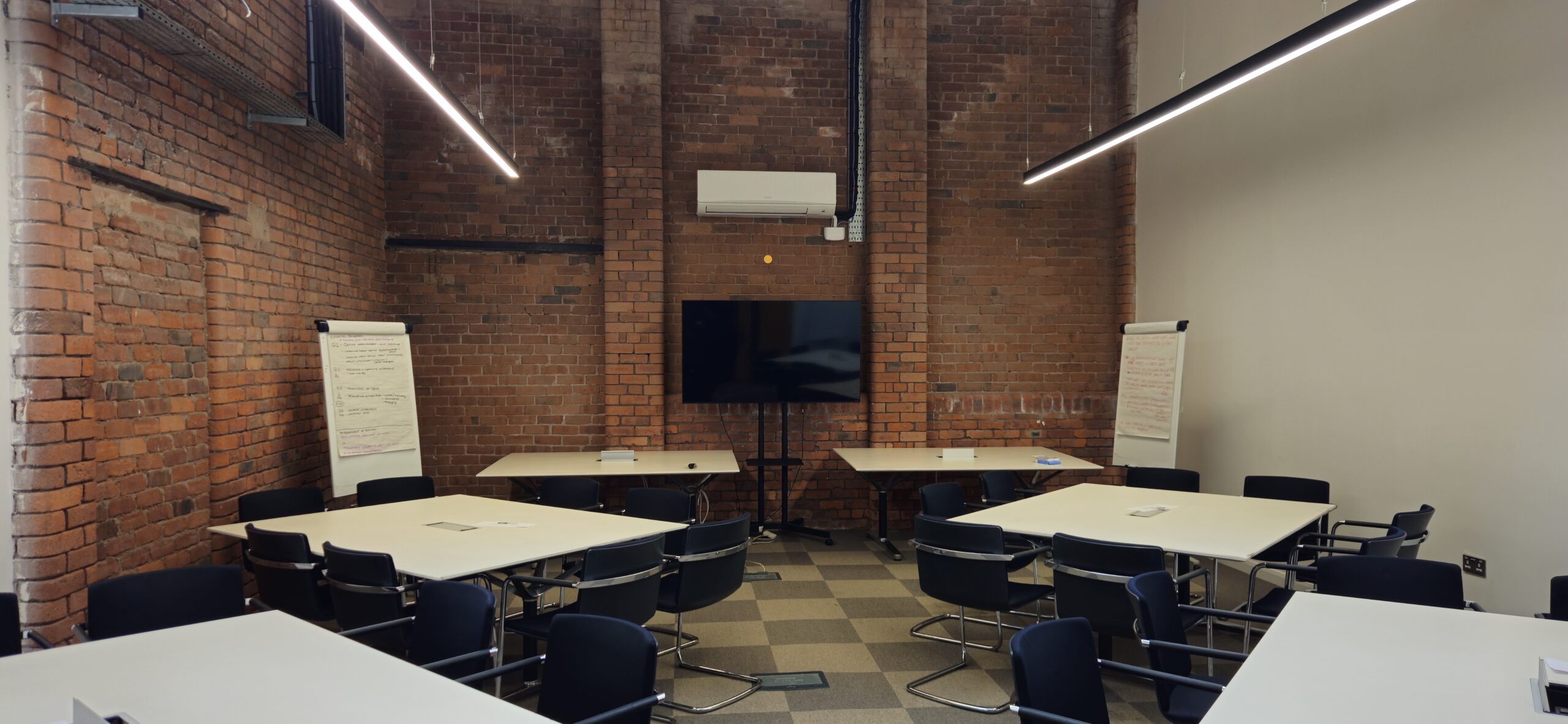 Meeting Room 2 professional meeting room at Ram Mill Business Centre in Oldham, featuring exposed brick walls, a large display screen, flipchart, and modular table setup. Perfect for team discussions, strategy sessions, and workshops.