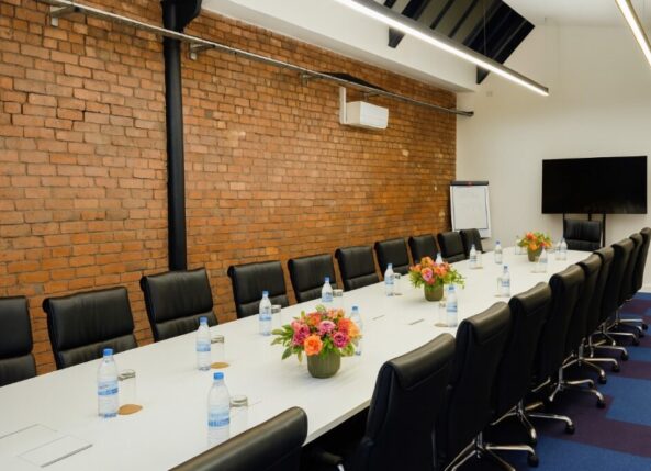 xecutive-style meeting room at Ram Mill Business Centre in Oldham, featuring a long conference table, sleek leather chairs, exposed brick walls, and a large screen for presentations. Ideal for board meetings, client pitches, and professional gatherings